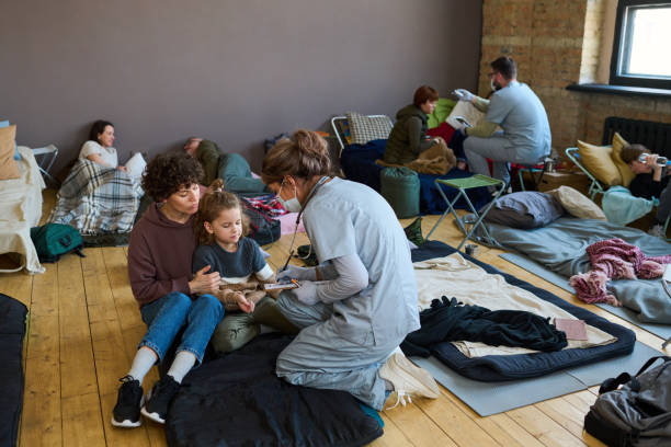 Healthcare worker giving medical help to young female refugee and little boy Healthcare worker in blue uniform, protective mask and gloves giving medical help to young female refugee and little boy in camp emergency shelter stock pictures, royalty-free photos & images