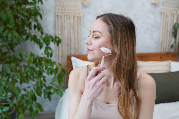 a jovem está fazendo massagem facial gua sha no banheiro. pedra de jade gua sha em tratamento domiciliar - lastone therapy massaging stone shiatsu - fotografias e filmes do acervo