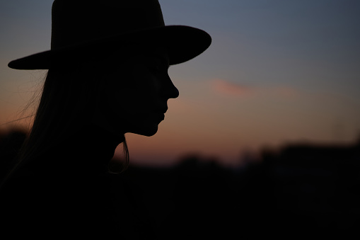 Closeup silhouette portrait of happy fashionable long hair woman wearing hat at sunset. Image against sun and blue sky. High quality horizontal photo