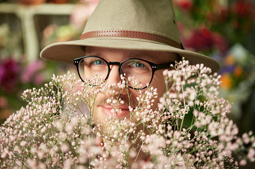 Portrait of cheerful male person wearing eyeglasses and hat in fresh flower bouquet. Fresh spring flowers. Mother's Day, Valentine's Day or International Women's Day concept. High quality photo
