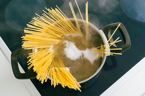 Spaghetti in pot is cooked in boiling water on electric ceramic hob. Top view, soft focus, yellow gluten free corn pasta cooking bolognese