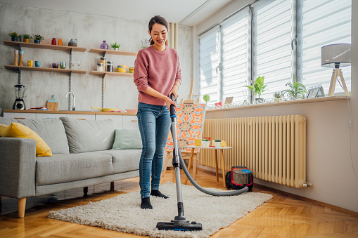 Young cheerful Japanese woman cleaning house with vacuum cleaner