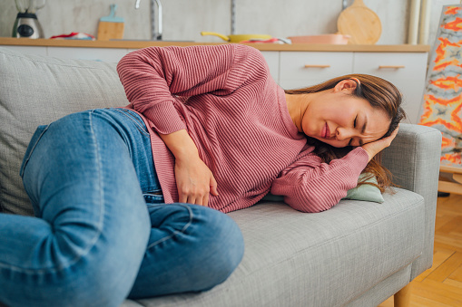 Young Japanese woman lying on her sofa alone and suffering from period cramps at home