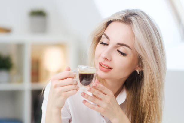 A young beautiful Caucasian blonde woman rests at home. A woman sits at a table, drinking coffee from a glass cup and reading a magazine. stock photo