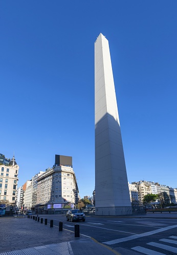Buenos Aires, Argentina, October 31, 2019: A car passes the Obelisco de Buenos Aires at sunrise. The obelisk is a national historic monument and icon of the city, in the morning sunlight. It was erected in 1936 to commemorate the quadricentennial of the first foundation of the city.