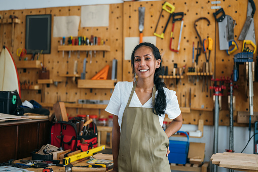 Positive woman working in carpenter workshop . Shot of smiling confident young Asian female joiner in apron standing near workbench and looking at camera friendly while working in craft workshop