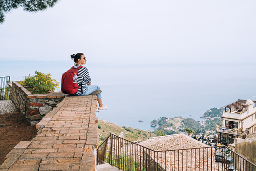 Tourist woman sitting with city backpack and enjoying ancient city harbor on the Ionian seacoast. Small Italian Castelmola Comune over the Taormina. Eastern Sicily, Italy.