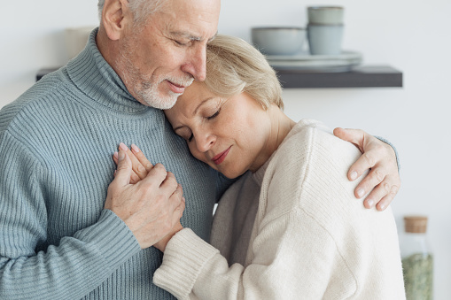 Concept of true love, sincere people, tenderness and romantic in relationships. Grey haired husband hug his calm and tranquil wife. Marriage couple cuddle to each other, standing at home