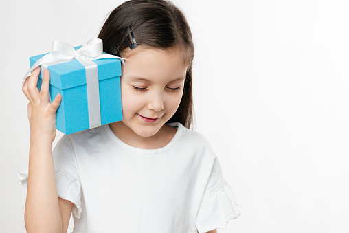 Front view of a cute little girl is holding a blue gift box in her hand with closed eyes trying to hear what is inside the gift box.