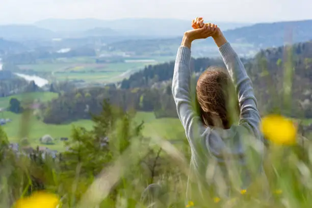 Young woman with rear view raising arms and making relaxing exercises with beautiful view from hill down to Aare River valley.