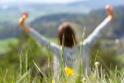 Blurred background with sitting young woman in spring meadow at rear view, raising arms and making relaxing exercises with beautiful view from hill to valley.