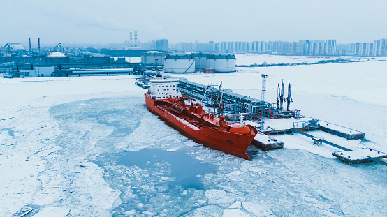 Northern oil terminal in snow and tanker illuminated by red lights moored on ocean water covered with ice pieces at evening twilight aerial view