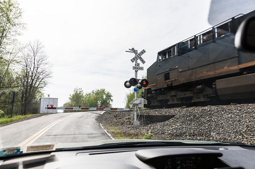 Car point of view with a low angle view looking uphill though the windshield of a stopped, waiting car at a grimy, fast moving, diesel electric locomotive freight train engine speeding through a dangerous rural road intersection railroad crossing at the crest of a \