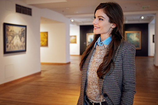 A young art dealer looking around at numerous fine art paintings hanging on the walls of a gallery