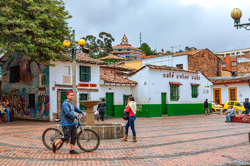 Bogota, Colombia - July 20, 2016: Local Colombian people and a few tourists are seen enjoying the small square called Chorro de Quevedo in the La Candelaria district of Bogotá, the Andean capital city of the South American country of Colombia. It is here that the Spanish Conquistador, Gonzalo Jiménez de Quesada founded the city in 1538. It is also from this Square that the Zipa, or Chief of the Muisca tribe, viewed the Sabana de Bogotá regularly. In 1832 the site was purchased by the Augustinian priest, Father Quevedo, who installed the public water fountain on the Square. Photo shot on an overcast morning; horizontal format.