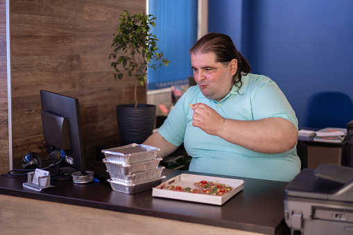 A large busy man is holding a slice of pizza in one hand and typing on a keyboard with his other for a busy lunch at the office, more containers of food are sitting on the desk