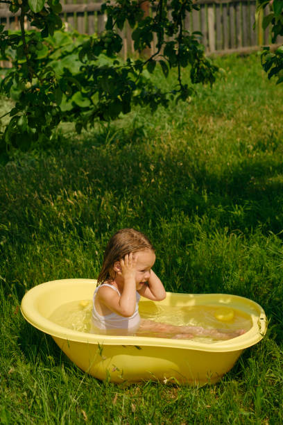 a wet little funny cute girl splashes in a bath in the garden in nature on a hot sunny day. the child takes a fresh bath - bird water bath garden stockfoto's en -beelden