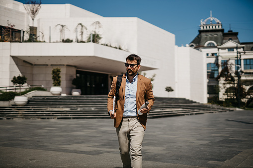 Spontaneous image of a confident looking, elegant-dressed, handsome bearded man walking outdoors, in a city, with a backpack, thermos and laptop in his hands. He is wearing stylish sunglasses and a suit.