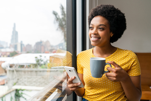 Portrait of a happy black woman at home drinking coffee while using an app on her phone and looking through the window - lifestyle concepts