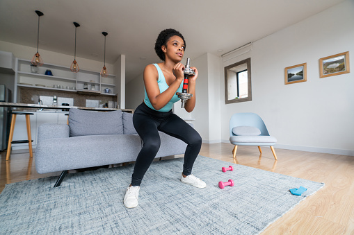 Beautiful African American woman exercising at home using dumbbells and squatting - healthy lifestyle concepts