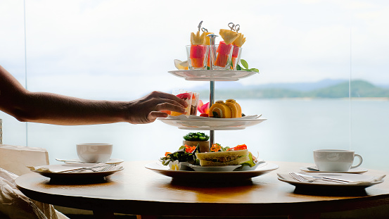 Woman hand takes piece of cake from afternoon tea set for two persons, fresh fruits on bamboo sticks skewers, cakes, pastries, sandwiches, snacks, tea cups in luxury hotel restaurant, sea background