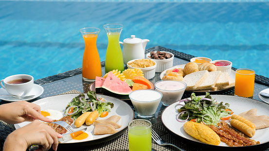 Vacation breakfast table. Woman hands cutting sunny side eggs at luxury restaurant or villa hotel room near the blue swimming pool on tropical holidays. Breakfast for two, romantic, honeymoon travel.