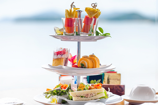 Traditional english high tea stand with selection of sweets, cakes, sandwiches, fruit canapes. Afternoon tea tray set with sweet treats on table. Tropical sea and mountains landscape background.