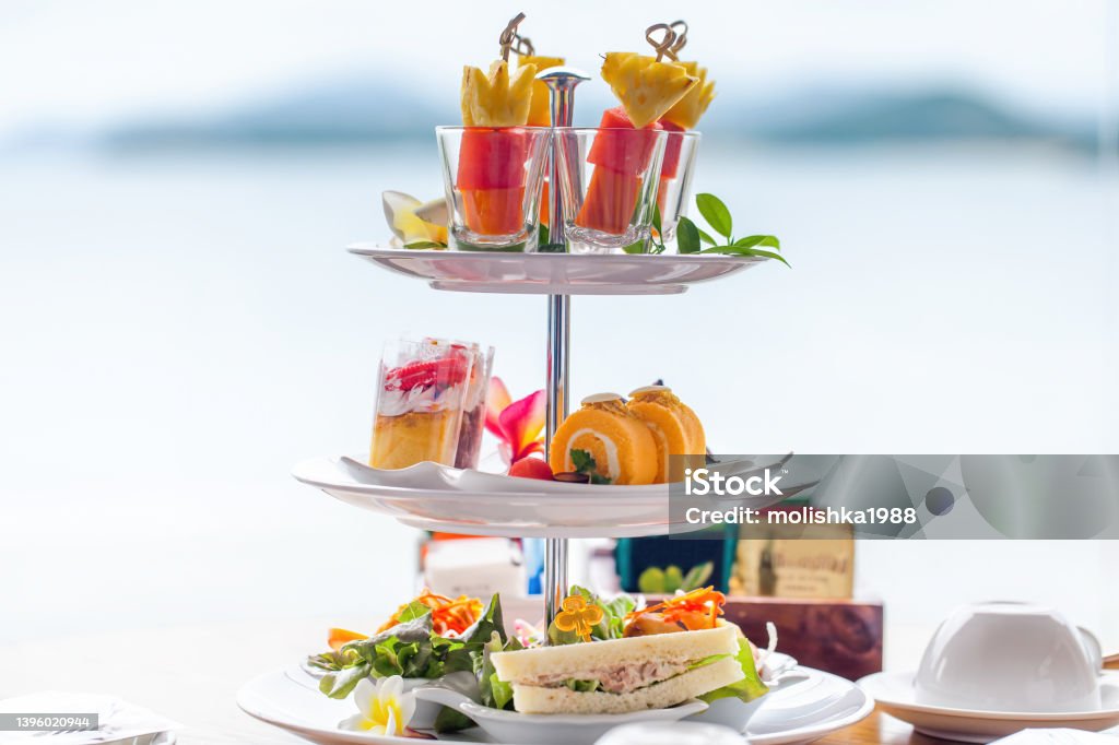 Traditional english high tea stand with selection of sweets, cakes, sandwiches Traditional english high tea stand with selection of sweets, cakes, sandwiches, fruit canapes. Afternoon tea tray set with sweet treats on table. Tropical sea and mountains landscape background. Afternoon Tea Stock Photo