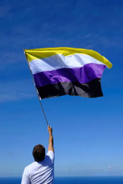 Caucasian man in white t-shirt waving non-binary flag in the wind over a radiant blue sky.