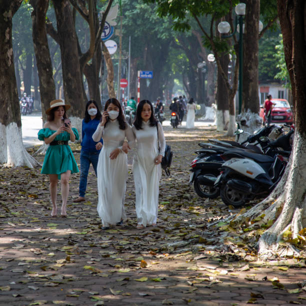 Hanoi, Vietnam - March 26, 2022: pedestrians, including two women in traditional Ao Dai dresses, walk along  Phan Dinh Phung street Hanoi , a tree-lined street known for elegant french style villas