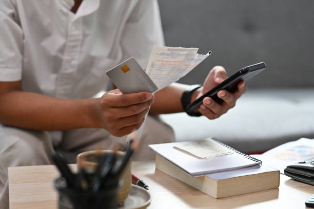 istock Close up with man sitting on a couch while holding bills and credit card and using mobile phone for calculate, finance, business, home and technology concept. 1396046862