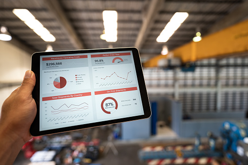 Action of manager is looking at logistic summary information on tablet with factory warehouse as background. Industrial and business investment concept photo. Close-up and selective focus.