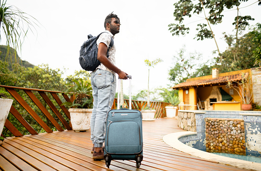 Young man pulling a wheeled suitcase along the wooden deck after arriving at his rental accommodation