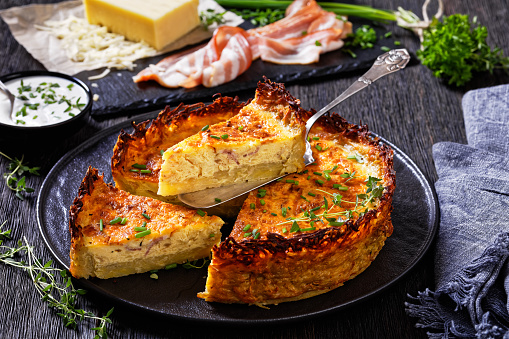 close-up of  bacon and cheddar quiche with hash brown crust on black plate, one piece on cake spatula. ingredients at background, horizontal view fro above