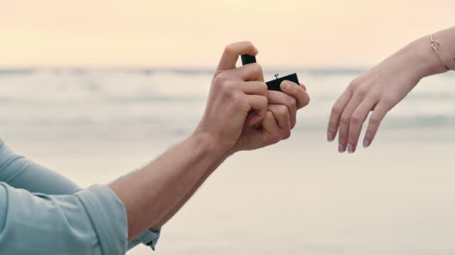An unrecognizable couple during their proposal at the beach. Closeup of a man placing an engagement ring on his girlfriend’s finger