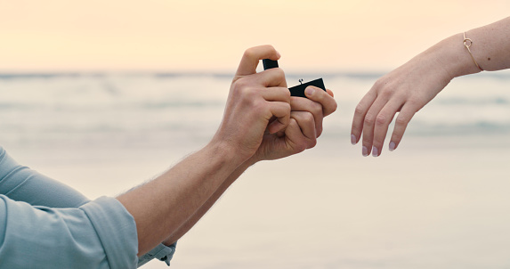An unrecognizable couple during their proposal at the beach. Closeup of a man placing an engagement ring on his girlfriend’s finger