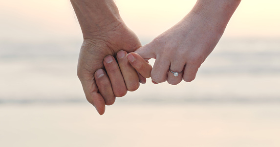 An unrecognizable couple holding hands and wearing an engagement ring. Closeup of a man and woman after their proposal at the beach
