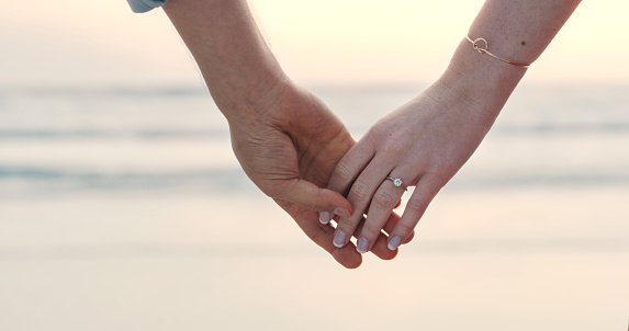 An unrecognizable couple holding hands and wearing an engagement ring. Closeup of a man and woman after their proposal at the beach