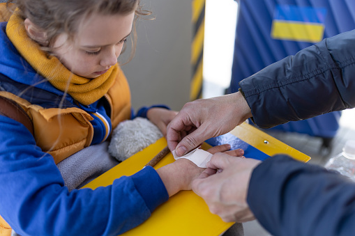 A volunteer helping Ukrainian refugee child at train station, giving plaster to injured hand.