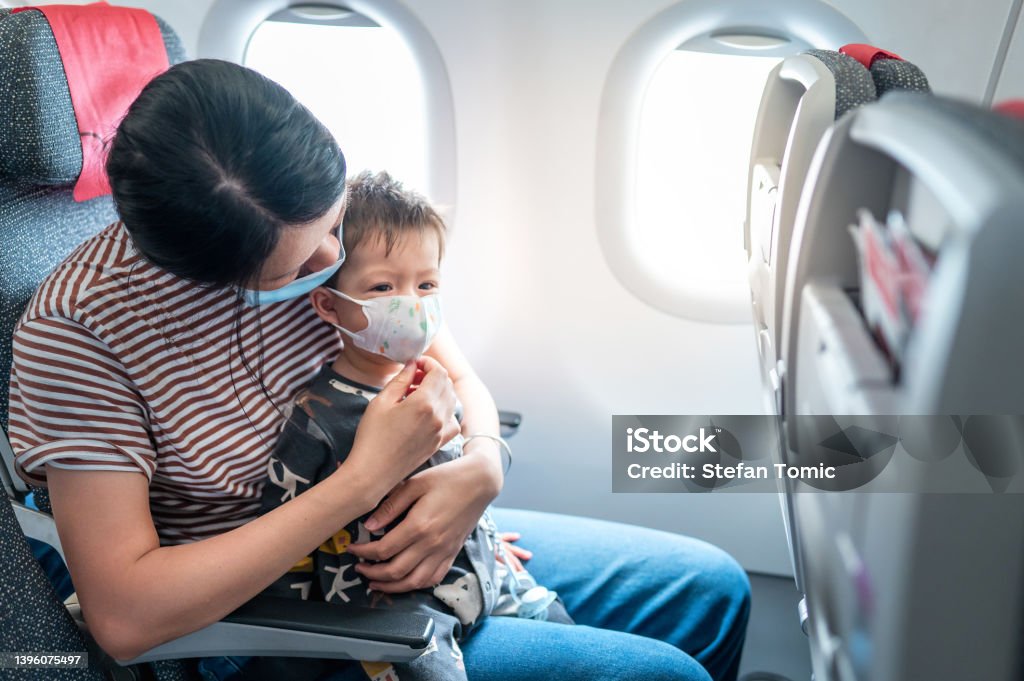Mother and her baby boy wearing surgical face mask in an airplane. Baby traveling in airplane flying sitting on his mother lap in the aircraft Mother and her baby boy wearing surgical face mask in an airplane. Baby traveling in airplane flying sitting on his mother lap in the aircraft. Infant traveling in airplane flying sitting on his mother lap in the aircraft while following Covid-19 procedures for corona virus prevention Post-Pandemic Stock Photo