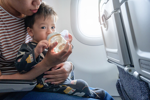 Mother taking airplane with a baby boy who is drinking water from his bottle. Infant traveling in airplane flying sitting on his mother lap in the aircraft