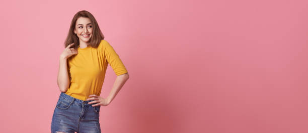 Pretty smiling woman in casual yellow t-shirt looking to copy space aside on pink studio background. stock photo