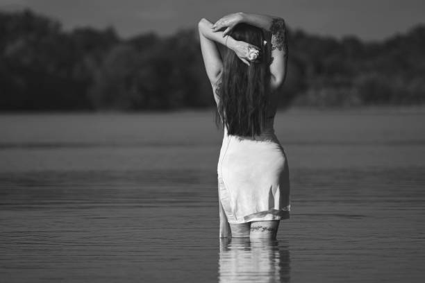 Black and white photo of a young woman with long dark hair, wearing white dress, turned backwards to camera, in a lake.