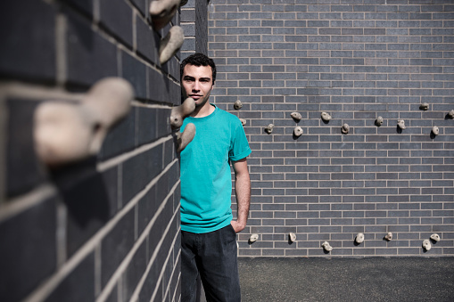 Young male posing in a boulder space in the city. He is standing and looking at camera. We can see grips on the brick wall.