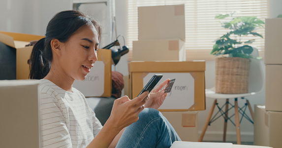 Young woman moving house sit on floor holding smartphone and banking credit card after unpacking.
