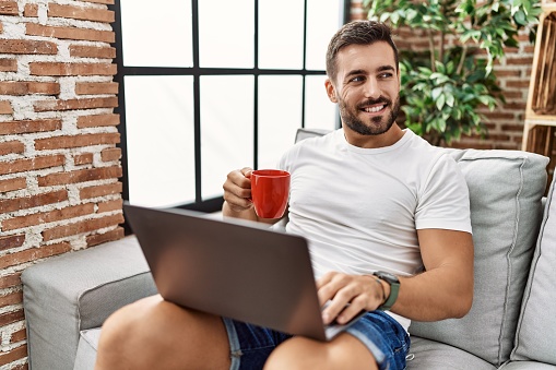 Young hispanic man using laptop and drinking coffee at home