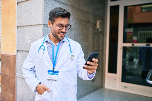 Young handsome hispanic doctor wearing uniform and stethoscope smiling happy Standing with smile on face using smartphone at town street.