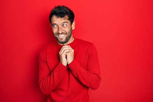 Handsome man with beard wearing casual red sweater laughing nervous and excited with hands on chin looking to the side