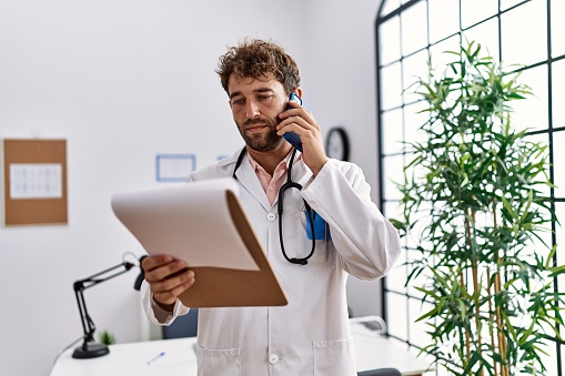 Young hispanic man wearing doctor uniform reading diagnosis talking on the smartphone at clinic