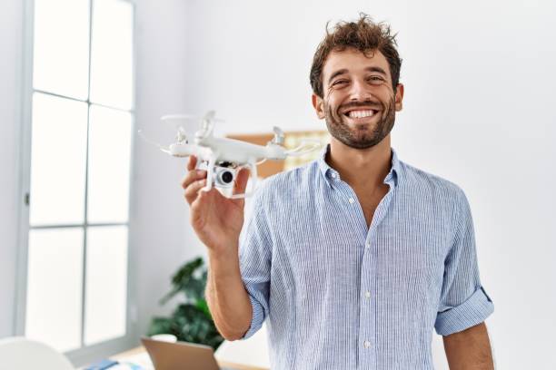 Young handsome man using drone at architect office looking positive and happy standing and smiling with a confident smile showing teeth stock photo